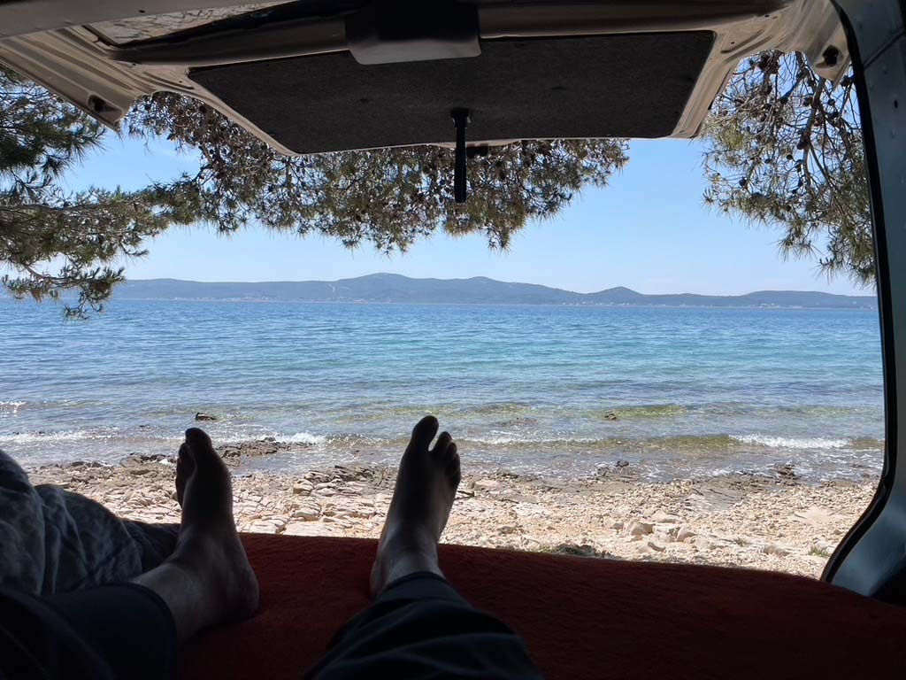 Blick aus dem geöffneten Heck eines Vans auf eine traumhafte Mittelmeer-Kulisse. Ein Reisender liegt entspannt auf einer roten Decke im Fahrzeug, die Beine ausgestreckt, während er auf das kristallklare, blau schimmernde Wasser und die fernen Bergketten am Horizont schaut. Über dem Van spenden dichte Pinienzweige Schatten und rahmen das Bild ein. Diese Szene fängt das Gefühl von Freiheit und Abenteuer perfekt ein – ein mobiles Zuhause direkt am Meer