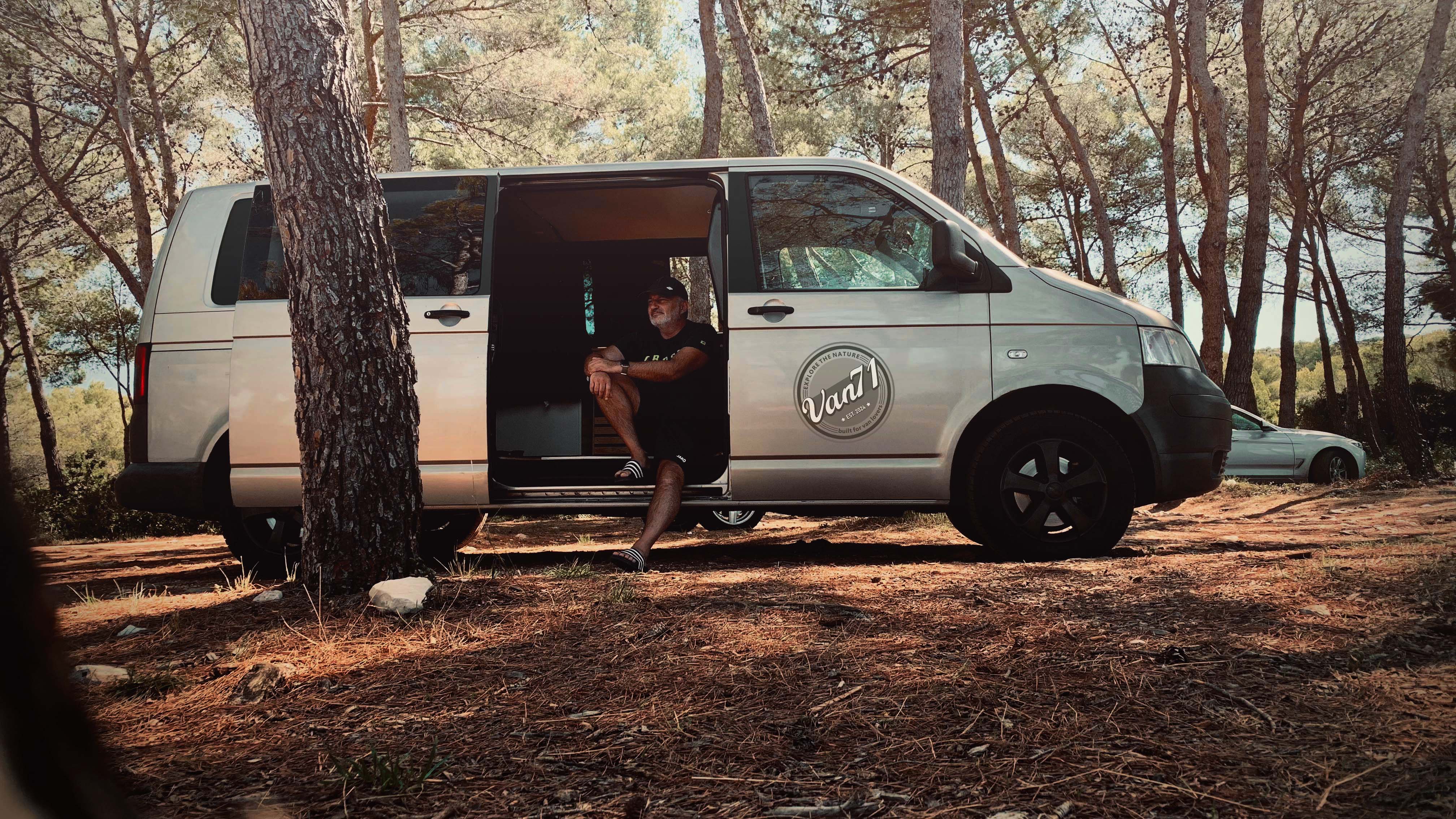 Ein Mann sitzt entspannt in der geöffneten Seitentür eines silbernen Vans mit Van71-Logo, umgeben von einem lichten Pinienwald. Das warme Licht der Sonne fällt durch die Bäume und erzeugt eine ruhige, naturverbundene Atmosphäre. Der Blick schweift in die Ferne – ein Moment der Zufriedenheit und Reflexion nach der erfolgreichen Umsetzung des ersten eigenen Innenausbaus